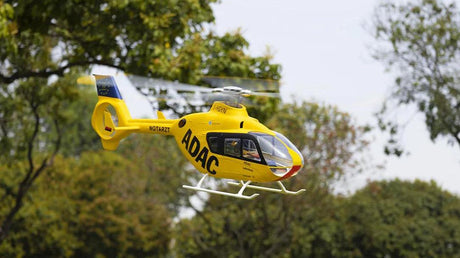 FLYWING EC135 470-scale RC helicopter in yellow ADAC livery flying outdoors, showcasing its realistic design and 810mm rotor against a backdrop of green trees